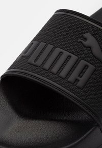 Sandale noire à glissière avec une sangle texturée arborant le logo « PUMA » embossé et une petite silhouette de chat. Semelle intérieure en caoutchouc lisse pour un confort optimal.