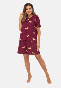 Kobieta stojąca boso, ubrana w burgundową bluzkę z krótkim rękawem i dopasowane szorty w żółte wzory w kokardki, pozująca z ręką przy włosach.