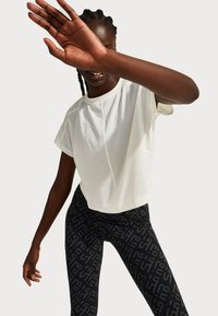 Person wearing a white short-sleeve top and black patterned leggings, raising one arm to partially cover their face, standing against a plain background.