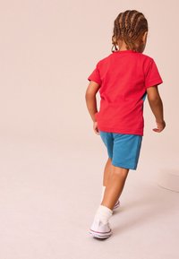 Rød kortærmet skjorte kombineret med blå shorts, begge lavet af bomuld. Barnet har hvide sneakers på og hvide op-rullede sokker.