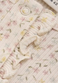 Licht geribbeld stoffen top in crème met pastelkleurig bloemenpatroon en gerafelde rand. Voorzien van een ronde knoop bij de halslijn. Zachte textuur.