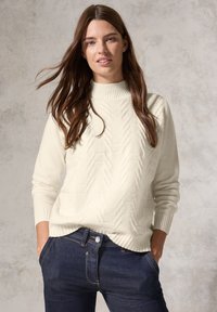 Maglione lavorato a maglia color crema con collo alto a coste e motivo a spina di pesce, abbinato a jeans in denim scuro. Texture morbida e vestibilità rilassata.