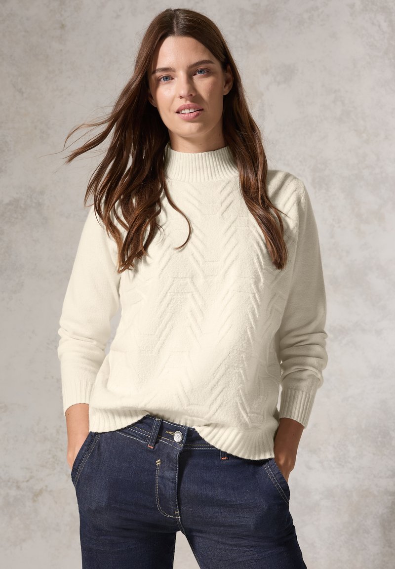 Maglione lavorato a maglia color crema con collo alto a coste e motivo a spina di pesce, abbinato a jeans in denim scuro. Texture morbida e vestibilità rilassata.