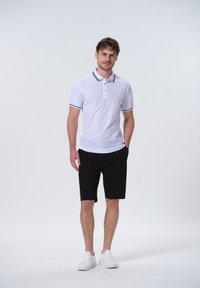 Hvid polo shirt med sort krave og ærmeaccenter, parret med sorte shorts og hvide sneakers. Modellen står foran en ensfarvet baggrund.