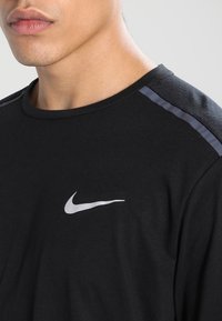 Schwarzes T-Shirt mit kurzen Ärmeln aus Baumwolle, mit einem silbernen Nike-Logo auf der Brust und strukturierten schwarzen Akzenten an den Schultern.