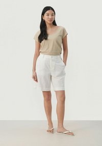 Beige kortärmad topp ihop med vita knä-långa shorts. Modell bär vita flip-flops och står på ett ljust, neutralt golv.