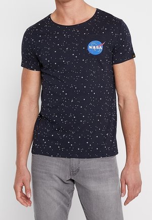 Mężczyzna w czarnej koszulce z białymi wzorami gwiazd i logo NASA na lewej piersi, w połączeniu z jasnoszarymi jeansami.