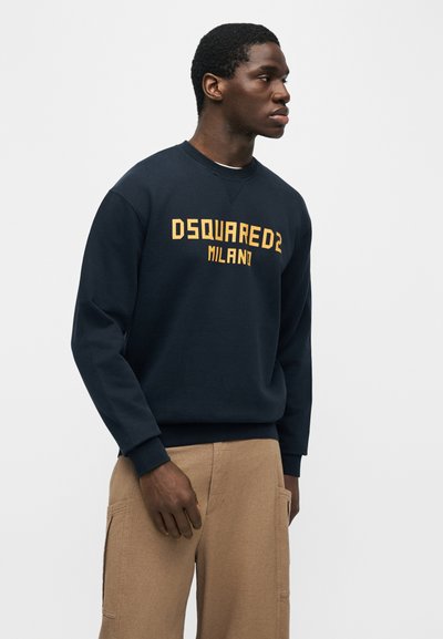 Νέος άνδρας που κοιτάζει προς τα δεξιά, φοράει μπλε σκούρο φούτερ με το κείμενο "DSQUARED2 MILANO" και μπεζ παντελόνι, στέκεται μπροστά σε ένα απλό φωτεινό φόντο.