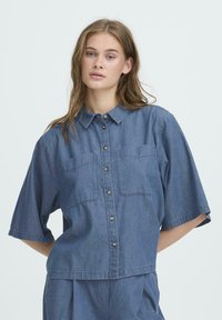 Denimhemd in mittlerem Blau, kurze Ärmel, zwei Brusttaschen, durchgehende Knopfleiste mit dunklen Knöpfen und lockerer Passform. Baumwollstoff.