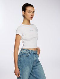 T-shirt bianca cropped con maniche corte che presenta la scritta azzurra "Grateful". Indossato con jeans in denim blu sbiadito a vita alta. Texture morbida.
