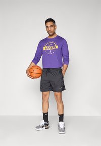 Lilla langærmet skjorte med "Lakers" logo, sorte shorts, Nike sneakers, holder en basketball. Glat stof, atletisk pasform.