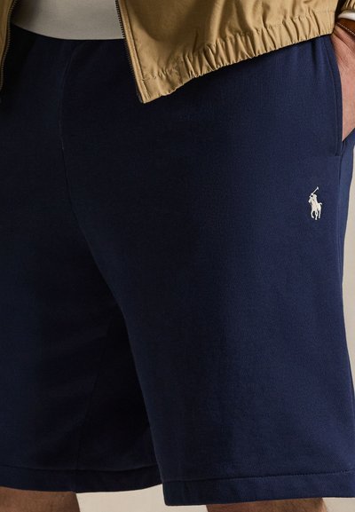 Man wearing navy blue shorts with a white embroidered polo player logo and a brown jacket, hand in pocket.
