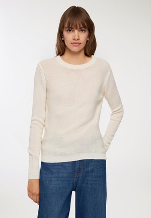 Giovane donna con capelli castani lisci che indossa un maglione a maglia color crema e jeans in denim blu, in piedi contro uno sfondo chiaro e semplice.