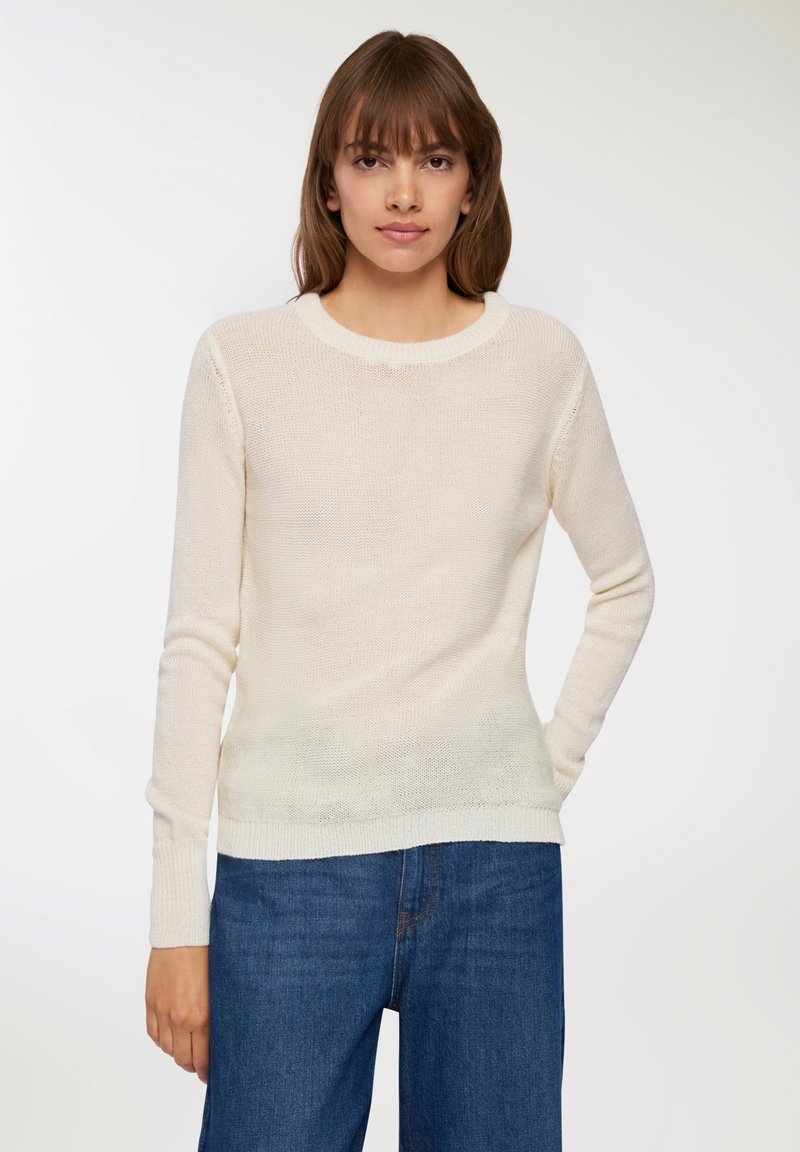 Giovane donna con capelli castani lisci che indossa un maglione a maglia color crema e jeans in denim blu, in piedi contro uno sfondo chiaro e semplice.