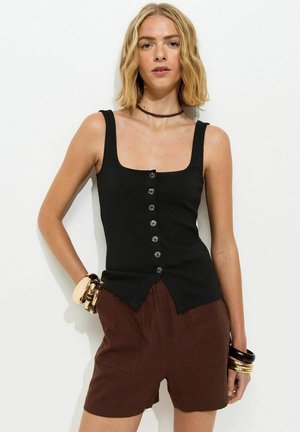 Vrouw met schouderlang blond haar draagt een mouwloos zwart topje met knopen, bruine korte broek en meerdere armbanden aan beide polsen.