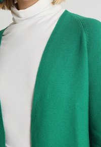 Grüne Strickjacke mit offenem Vorderteil, kombiniert mit einem weißen Rollkragenpullover. Die Struktur ist glatt und von mittlerer Dicke.