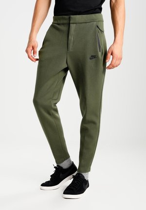 Mężczyzna noszący oliwkowo-zielone spodnie dresowe Nike z kieszenią na boku na zamek błyskawiczny, czarne trampki i szare skarpetki, stojący na białym tle.