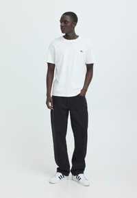 Witte katoenen t-shirt met ronde hals, met een klein zwart ooglogo op de borst. Gecombineerd met losse zwarte spijkerbroeken en witte sneakers.