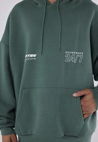 Πράσινη hoodie με μπροστινή τσέπη, κουκούλα με κορδόνι και εκτυπωμένο κείμενο "SOUNWAVE 24/7" και "RTRN". Φτιαγμένη από μαλακό, υφασμάτινο υλικό.