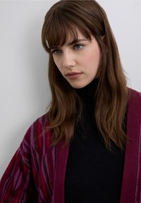 Cardigan featuring a burgundy and purple striped pattern with metallic accents, worn over a black turtleneck. Smooth texture and loose fit.