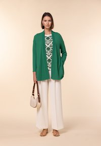 Grön cardigan, vit blommönstrad topp, vita vida byxor, guldsandaler och en beige handväska. Släta texturer, modern design.