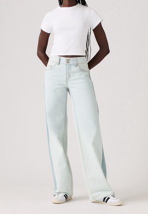 Persona con vaqueros de lavado claro y pierna ancha, camiseta blanca corta y zapatillas blancas con rayas negras, de pie con las manos detrás de la espalda.