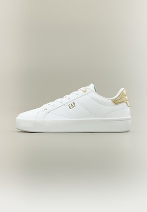 Valge madala lõikega toss kuldse kannaaktsendiga, valgete paelte ja "GAP" logoga küljel, esitatud ühevärvilise neutraalse tausta ees.