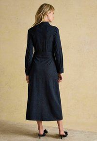 Dlhý rukáv, tmavomodré šaty s jemným leopardím vzorom, s opaskom v oblasti pásu a splývavou sukňou. Čierne muly na podpätku s detailmi v tvare mašlí.