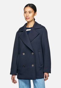 Cappotto doppiopetto blu navy con ampi rever, tasche frontali e dettagli in ottone. Realizzato in materiale testurizzato. Il modello indossa una maglietta a righe sotto.