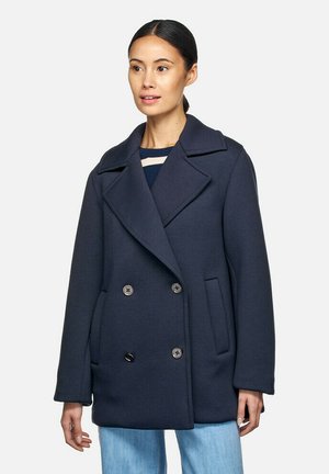 Cappotto doppiopetto blu navy con ampi rever, tasche frontali e dettagli in ottone. Realizzato in materiale testurizzato. Il modello indossa una maglietta a righe sotto.