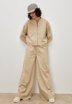 Donna con capelli ricci che indossa una giacca beige con zip, pantaloni larghi, berretto a scacchi e sneakers bianche, in piedi di fronte a un muro semplice.