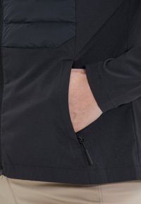 Giacca nera con zip frontale, dotata di un pannello toracico imbottito, tasche laterali e una superficie liscia. Design aderente, tessuto leggero.