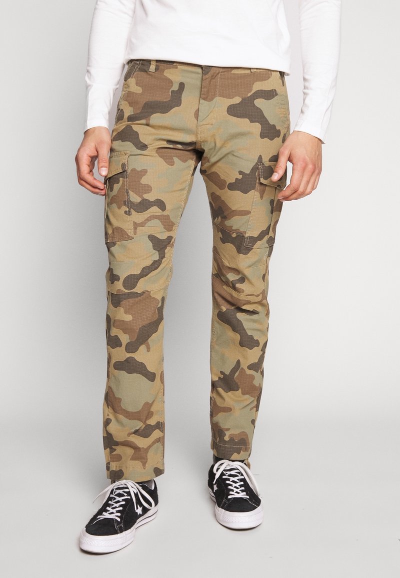 Pantalones cargo de camuflaje en tonos tierra con múltiples bolsillos, diseño de pierna recta y un acabado de tejido texturizado. Se llevan zapatillas negras.