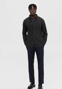 Camisa negra de botones con un acabado texturizado, combinada con unos pantalones de cuadros oscuros. El modelo lleva zapatos negros elegantes.