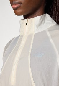 Veste de sport à demi-zip en beige clair. Comprend un tissu texturé, une marque discrète et un col montant. Fini lisse avec un détail de fermeture éclair.