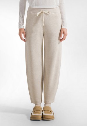 Beige, strukturierte Jogginghose mit einem Kordelzugbund, entspannter Passform und gerippten Bündchen. Kombiniert mit hellbraunen, gefütterten Hausschuhen und hellen Socken.