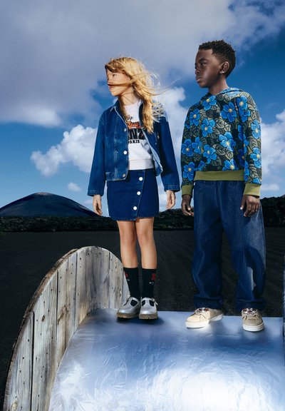 Giacca di jeans e gonna su una ragazza, felpa blu con fiori e pantaloni larghi su un ragazzo, entrambi su una superficie metallica, contro un cielo drammatico.