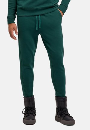 Uomo che indossa pantaloni jogger verde scuro e sneakers alte nere, in piedi con una mano in tasca e l'altra lungo il fianco.