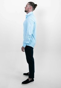 Lichtblauw button-up overhemd met lange mouwen, gedragen met donkere broeken en zwarte instappers; weergegeven in een zijprofiel tegen een witte achtergrond.