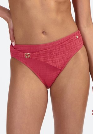 Donna che indossa slip bikini rosa testurizzati a vita alta con decorazioni in metallo color oro sulla parte anteriore e laterale.