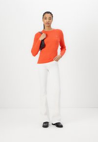 Suéter de punto de cable naranja con mangas largas, combinado con jeans blancos de campana y mocasines negros, de pie sobre un fondo liso.