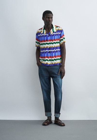 Άνδρας που στέκεται φορώντας πολύχρωμο πουκάμισο με γεωμετρικά σχέδια και κοντά μανίκια, γαλάζια τζιν με γυρισμένα μπατζάκια και καφέ σανδάλια σε μονόχρωμο φόντο.