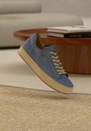 Blå semsket joggesko med beige snøring, vevd jute midt-sole, rund tå og bakre flik; hengt over et teksturert teppe.