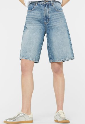Person trägt hellblaue, knieumspielte Jeans-Shorts und silberne Sneakers mit weißen Schnürsenkeln und steht vor einem einfarbigen Hintergrund.