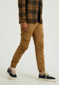 Tan cargo-byxor med flera sidofickor och en tapered ben-design, kombinerat med marinblå sneakers och en brun rutig jacka.