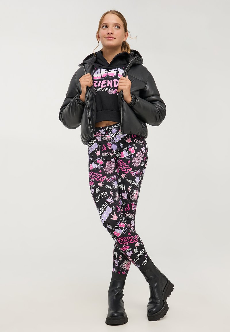 Giacca a vento nera con cappuccio, felpa nera corta con testo rosa "BEST FRIENDS" e leggings vivaci con motivi grafici colorati.