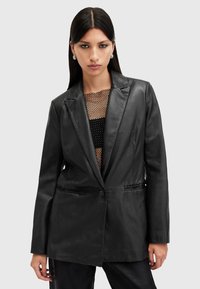 Černý kožený blazer se strukturovanými klopami, uzavírání na jeden knoflík a hladkým povrchem, nošený přes síťovaný top. Obsahuje dvě boční kapsy.