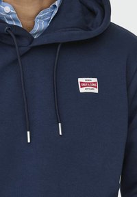 Dunkelblaue Kapuzenjacke mit einer Fronttasche, Kordelzügen und silbernen Spitzen. Mit einem weißen und roten Markenpatch auf der Brust. Weiche Textur.