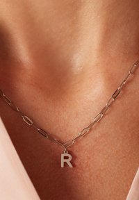 Zilveren metalen ketting met verlengde schakels, voorzien van een kleine "r" hanger. Glad textuur en minimalistisch ontwerp.