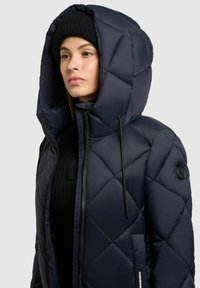 Jachetă puffer cu captuseală matlasată de culoare bleumarin, cu glugă, prezentând un fermoar frontal, șnur de strângere și detalii cu buton logo. Purtați peste un pulover negru cu dungi.